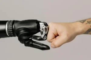 Obraz przedstawia zbliżenie na ludzką pięść uderzającą w dłoń robota. Mechaniczna ręka wygląda jak proteza, złożona z czarnych i metalicznych elementów. Na ludzkim przedramieniu widoczny jest tatuaż.