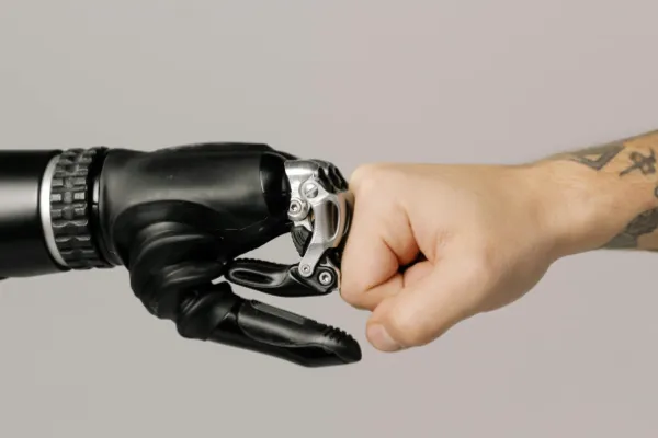 Obraz przedstawia zbliżenie na ludzką pięść uderzającą w dłoń robota. Mechaniczna ręka wygląda jak proteza, złożona z czarnych i metalicznych elementów. Na ludzkim przedramieniu widoczny jest tatuaż.